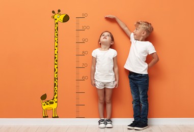 Image of Cute little children measuring their heights near orange wall with scale and drawing of giraffe