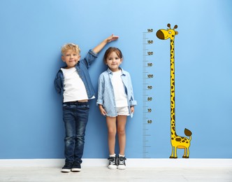 Image of Cute little children measuring their heights near blue wall with scale and drawing of giraffe