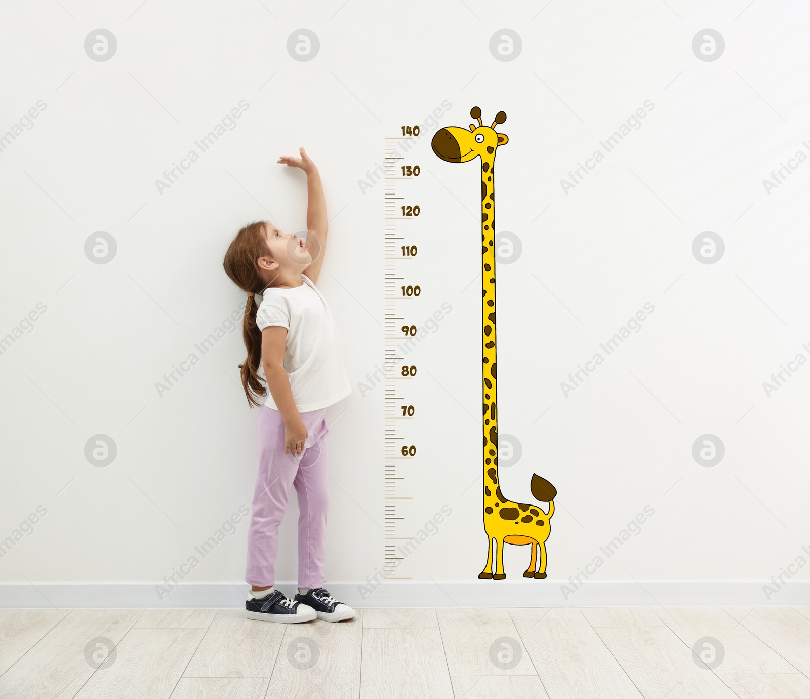 Image of Cute little girl measuring her height near white wall with scale and drawing of giraffe