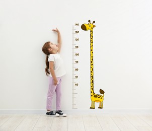 Image of Cute little girl measuring her height near white wall with scale and drawing of giraffe