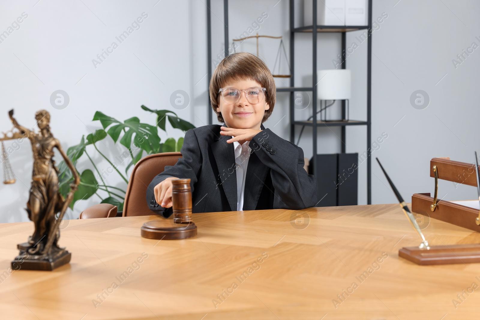 Photo of Choice of profession. Little boy in glasses with judge's gavel and statue of Lady Justice pretending to be lawyer at wooden table in office