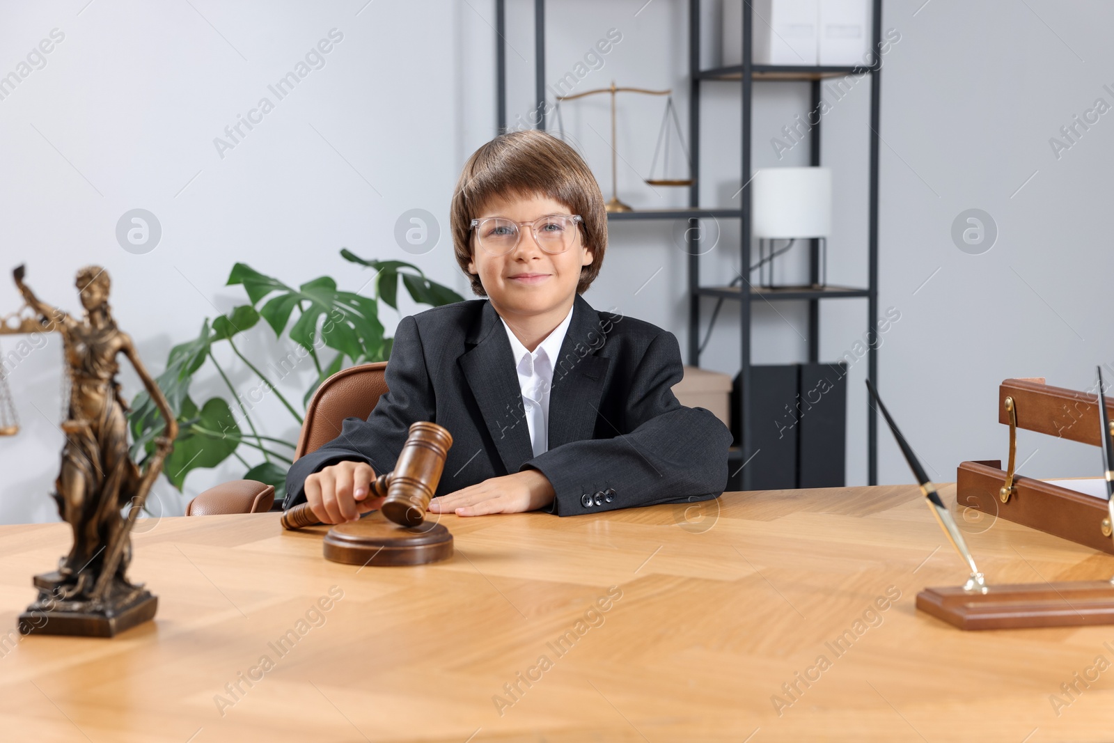 Photo of Choice of profession. Little boy in glasses with judge's gavel and statue of Lady Justice pretending to be lawyer at wooden table in office