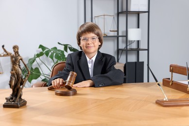 Photo of Choice of profession. Little boy in glasses with judge's gavel and statue of Lady Justice pretending to be lawyer at wooden table in office