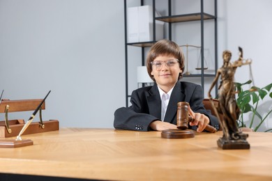 Photo of Choice of profession. Little boy in glasses with judge's gavel and statue of Lady Justice pretending to be lawyer at wooden table in office