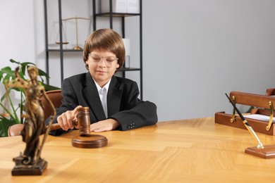 Photo of Choice of profession. Little boy in glasses with judge's gavel and statue of Lady Justice pretending to be lawyer at wooden table in office