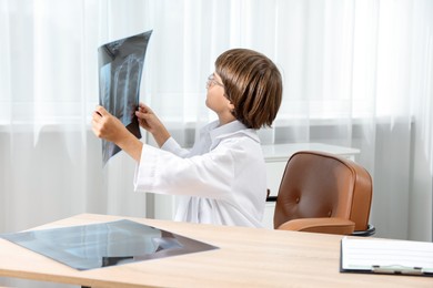 Photo of Choice of profession. Little boy in glasses and uniform with lungs x-ray pretending to be doctor at table in clinic