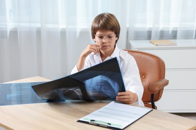 Photo of Choice of profession. Little boy in glasses and uniform with lungs x-ray pretending to be doctor at table in clinic