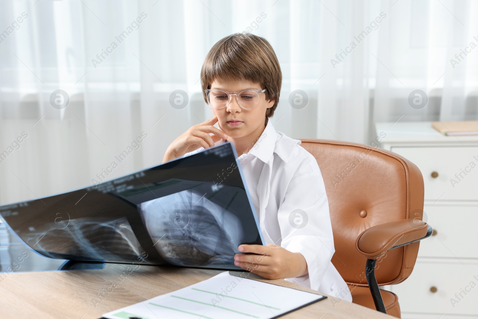 Photo of Choice of profession. Little boy in glasses and uniform with lungs x-ray pretending to be doctor at table in clinic