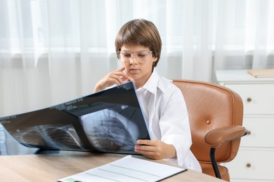 Photo of Choice of profession. Little boy in glasses and uniform with lungs x-ray pretending to be doctor at table in clinic