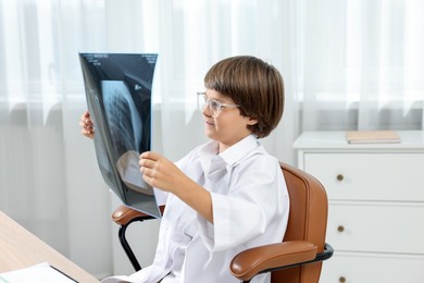 Photo of Choice of profession. Little boy in glasses and uniform with lungs x-ray pretending to be doctor at table in clinic