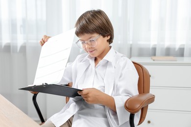 Photo of Choice of profession. Little boy in glasses and uniform with patient's medical card pretending to be doctor on chair in clinic