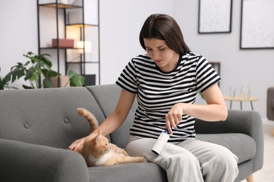 Photo of Woman with lint roller removing pet's hair from clothes and her cat on sofa at home