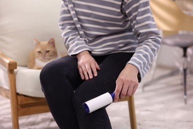 Photo of Woman with lint roller removing pet's hair from pants and her cat on armchair at home, closeup