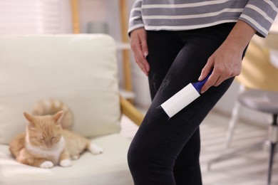 Photo of Woman with lint roller removing pet's hair from pants and her cat on armchair at home, closeup