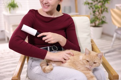 Photo of Smiling woman with lint roller removing pet's hair from clothes and her cat on armchair at home, closeup