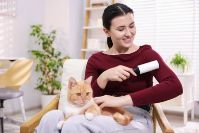 Photo of Smiling woman with lint roller removing pet's hair from clothes and her cat on armchair at home