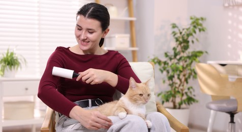 Photo of Smiling woman with lint roller removing pet's hair from clothes and her cat on armchair at home