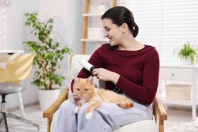 Photo of Smiling woman with lint roller removing pet's hair from clothes and her cat on armchair at home