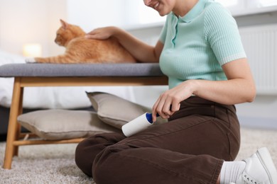 Photo of Smiling woman with lint roller removing pet's hair from pants and her cat on bench at home, closeup