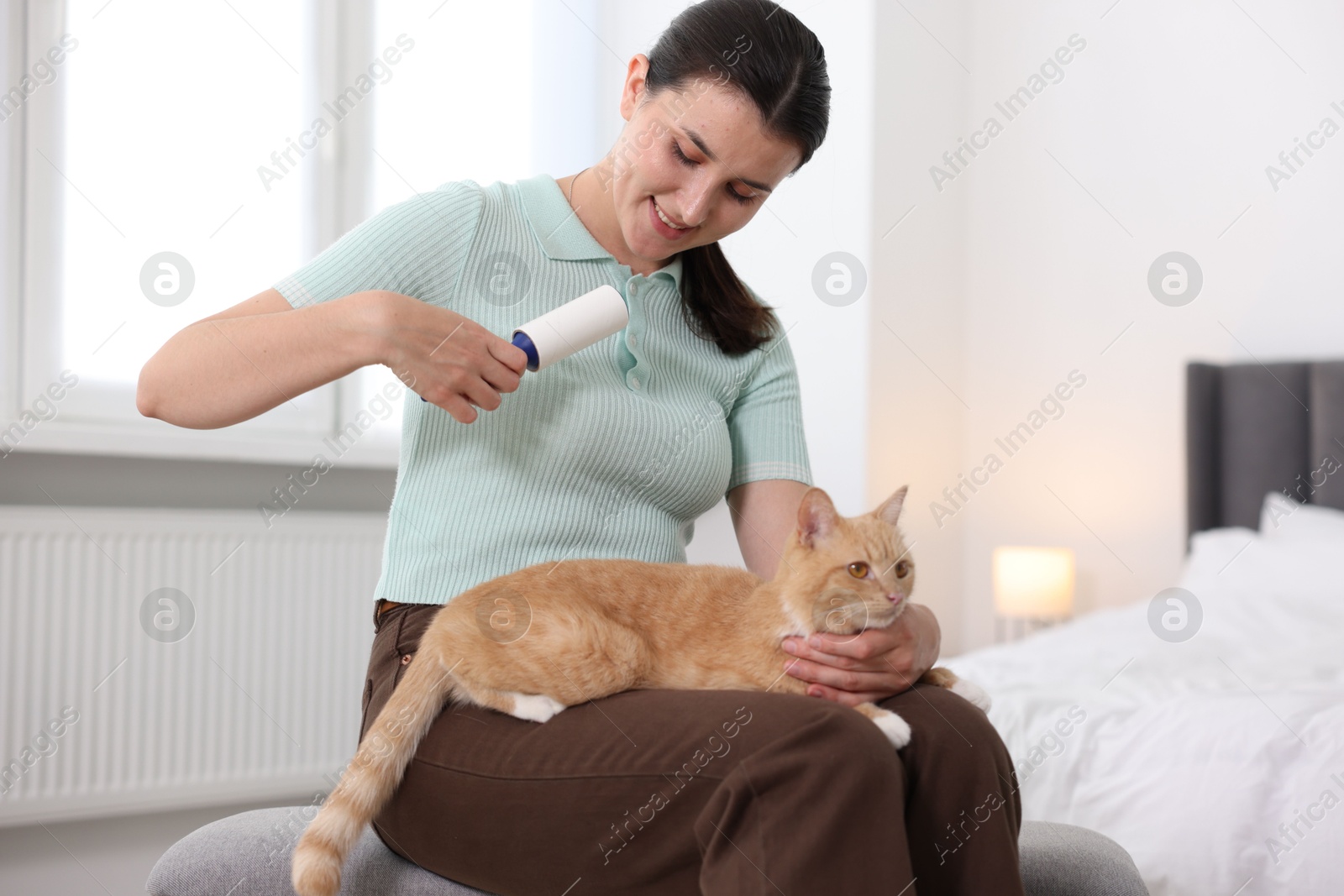 Photo of Smiling woman with lint roller removing pet's hair from clothes and her cat on bench in bedroom