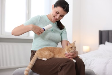 Photo of Smiling woman with lint roller removing pet's hair from clothes and her cat on bench in bedroom
