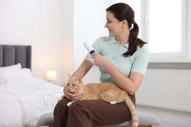 Photo of Smiling woman with lint roller removing pet's hair from clothes and her cat on bench in bedroom