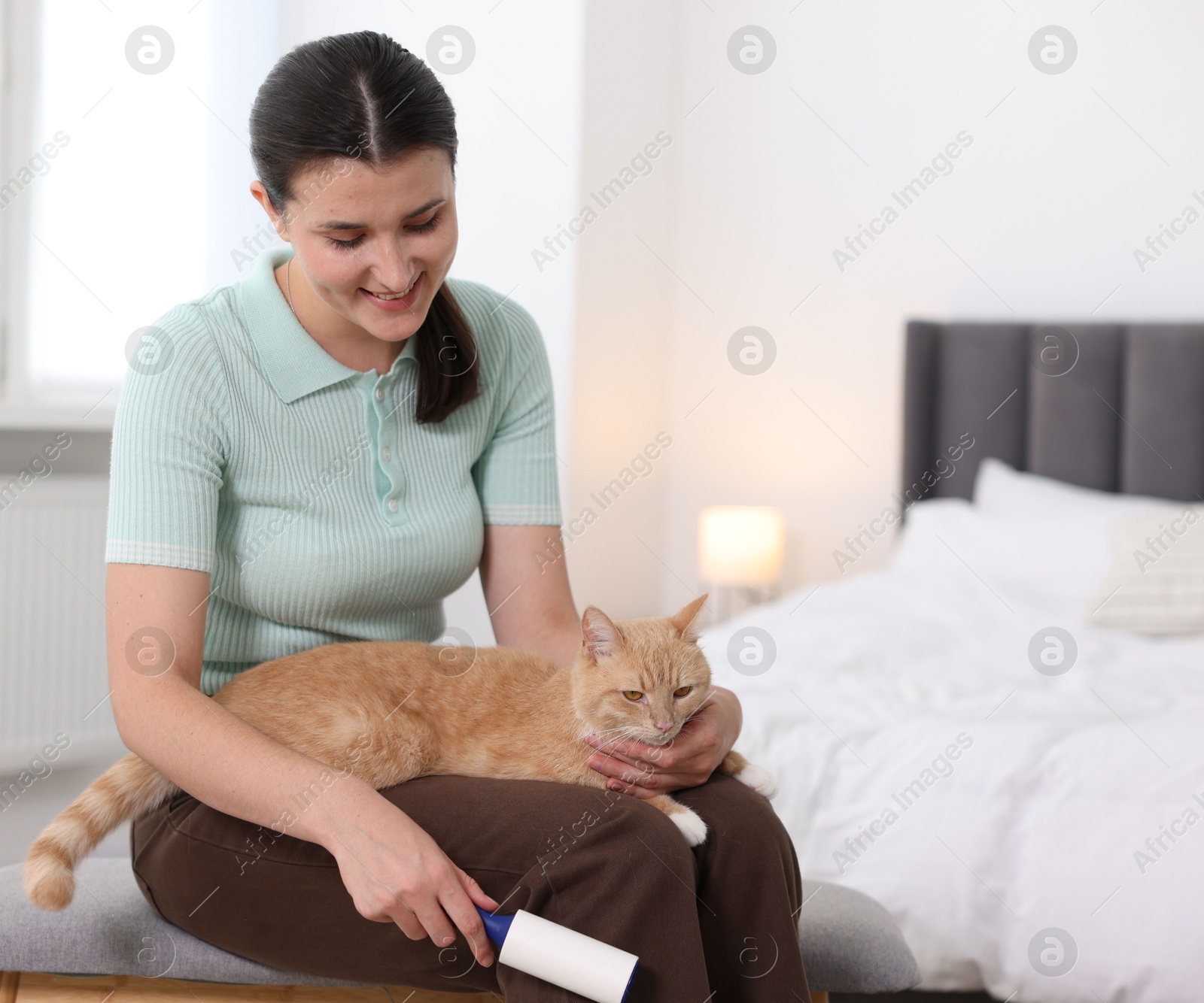 Photo of Smiling woman with lint roller and her cat on bench in bedroom. Space for text