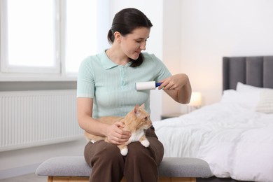 Photo of Woman with lint roller removing pet's hair from clothes and her cat on bench in bedroom