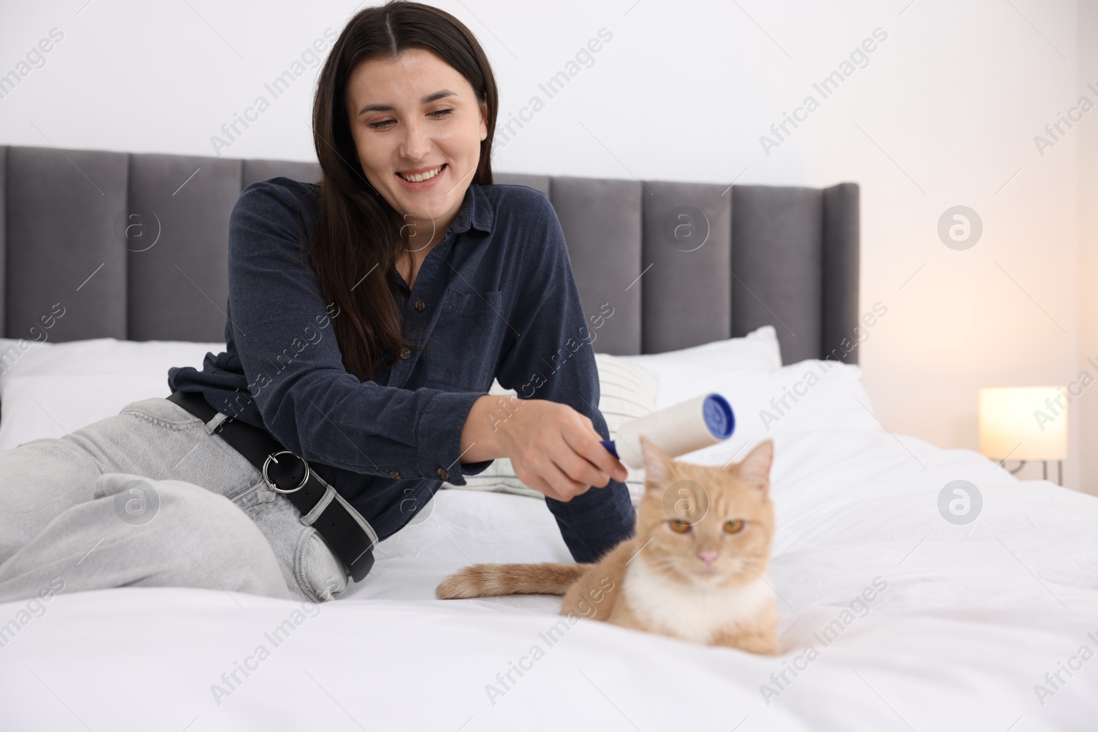 Photo of Smiling woman rubbing down her cat with lint roller on bed at home