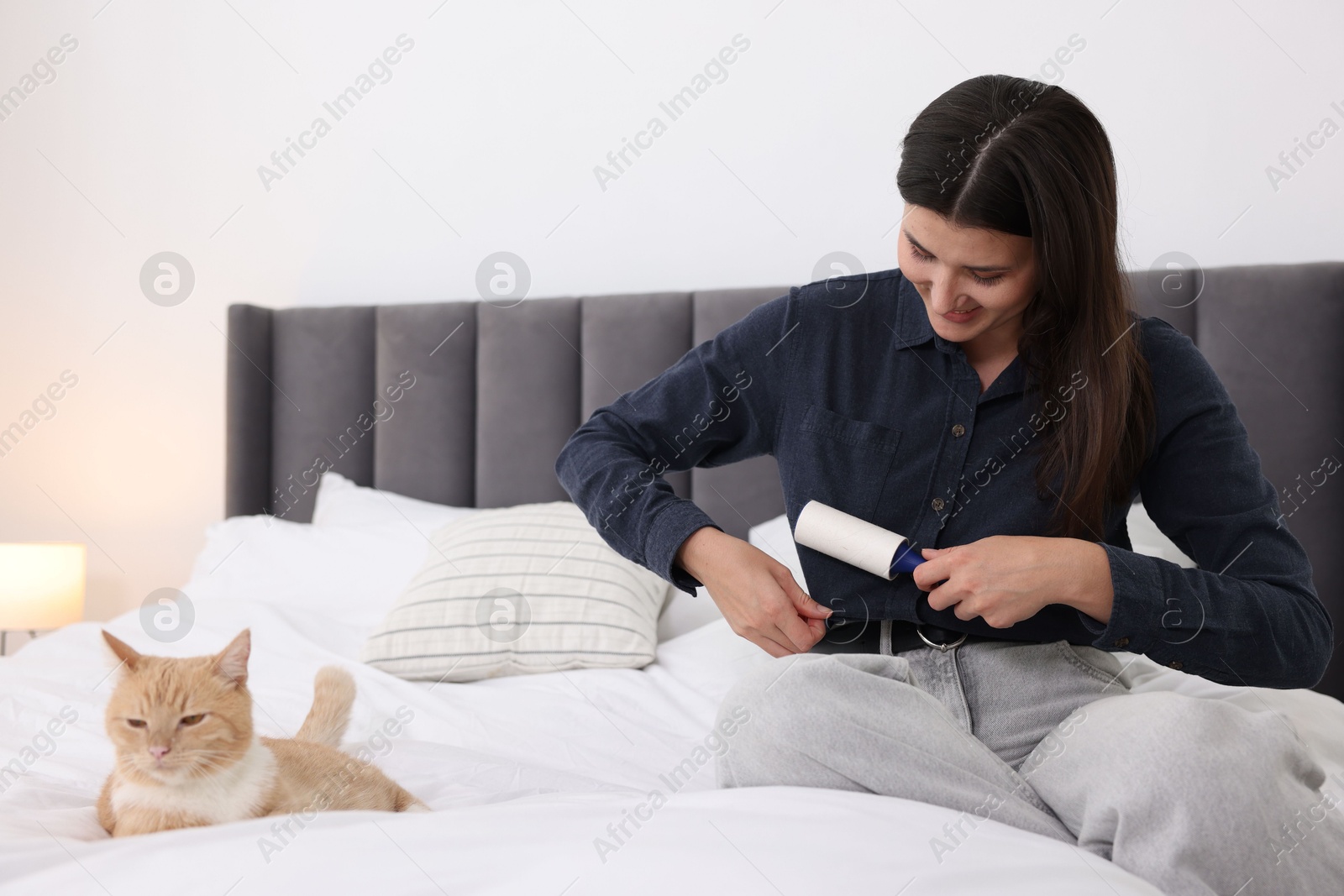 Photo of Woman with lint roller removing pet's hair from clothes and her cat on bed in bedroom