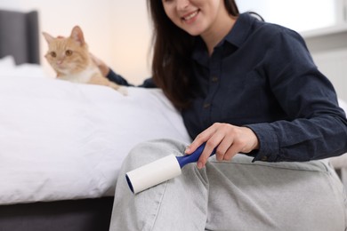 Photo of Smiling woman with lint roller removing pet's hair from clothes and her cat on bed at home, closeup