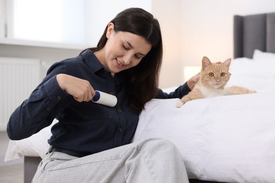 Photo of Smiling woman with lint roller removing pet's hair from clothes and her cat on bed at home