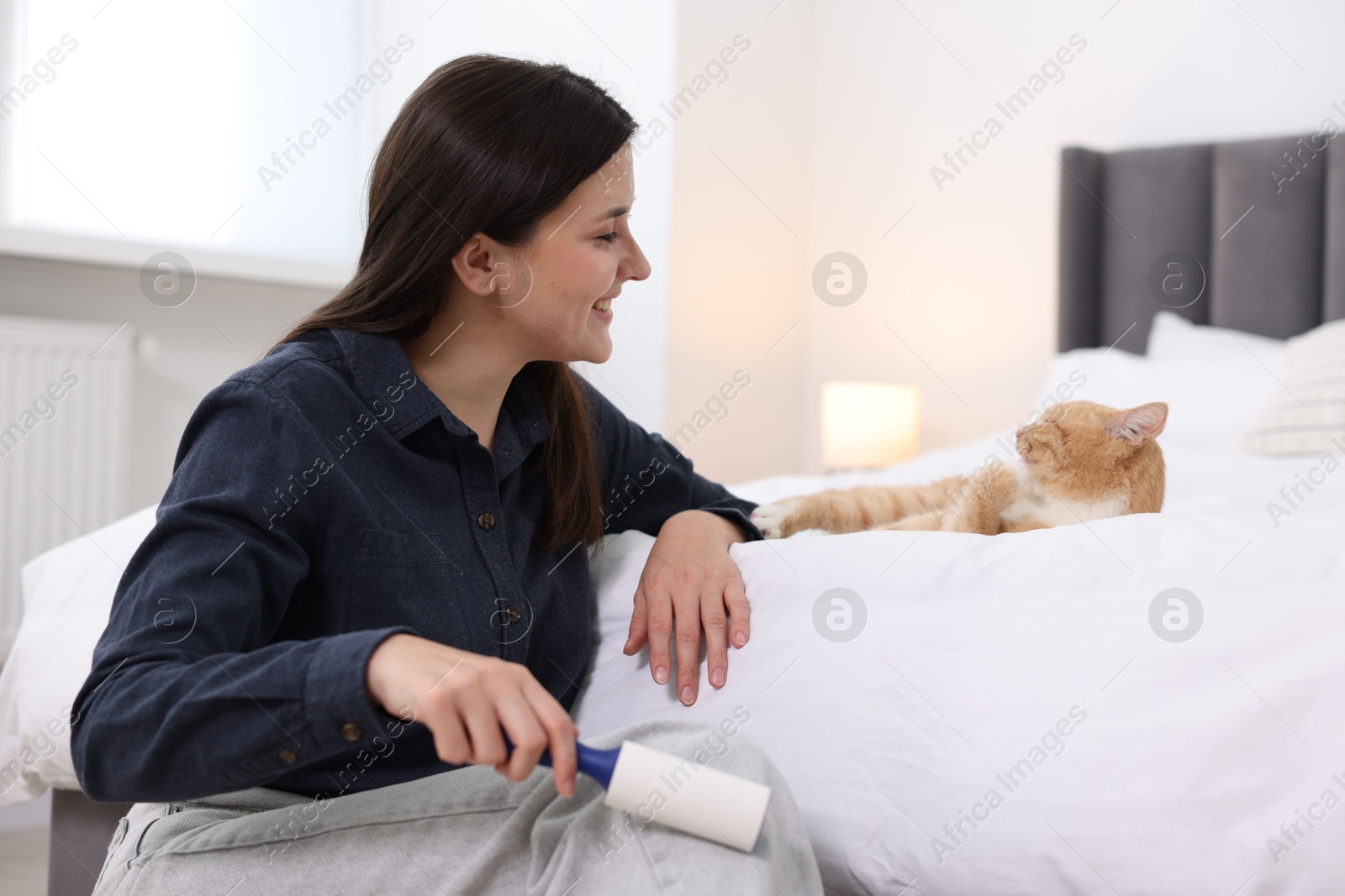 Photo of Smiling woman with lint roller removing pet's hair from clothes and her cat on bed at home