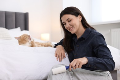 Photo of Smiling woman with lint roller removing pet's hair from clothes and her cat on bed at home