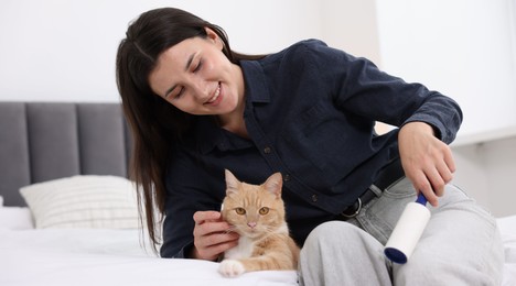 Photo of Smiling woman with lint roller removing pet's hair from clothes and her cat on bed at home