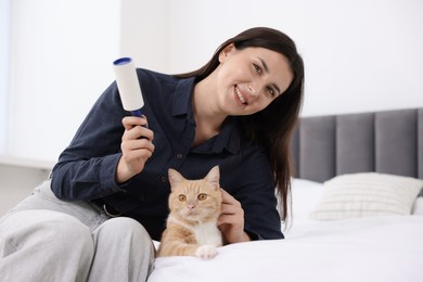 Photo of Smiling woman with lint roller removing pet's hair from clothes and her cat on bed at home
