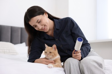 Photo of Smiling woman with lint roller removing pet's hair from clothes and her cat on bed at home