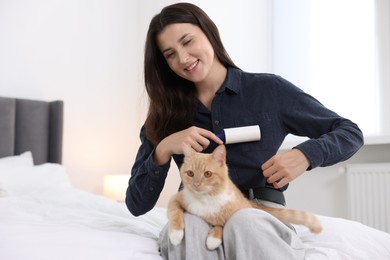 Photo of Smiling woman with lint roller removing pet's hair from clothes and her cat on bed at home