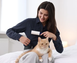 Photo of Smiling woman with lint roller removing pet's hair from clothes and her cat on bed at home