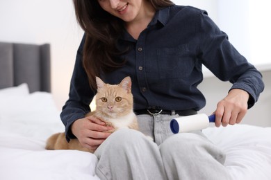 Photo of Smiling woman with lint roller removing pet's hair from clothes and her cat on bed at home, closeup