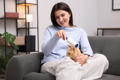 Photo of Smiling woman rubbing down her cat with lint roller on sofa at home