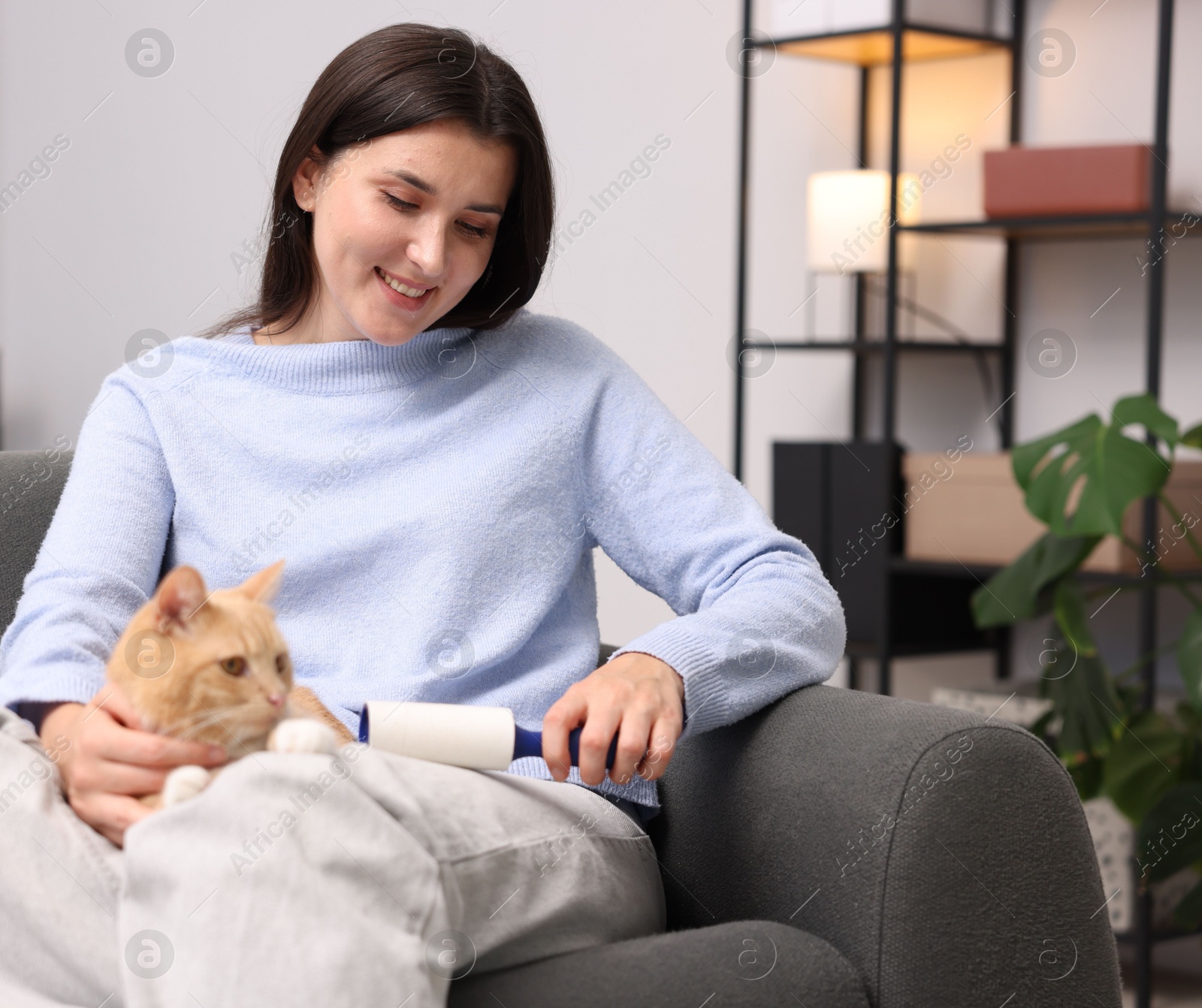 Photo of Smiling woman with lint roller removing pet's hair from clothes and her cat on sofa at home