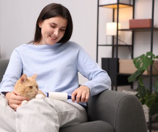Photo of Smiling woman with lint roller removing pet's hair from clothes and her cat on sofa at home
