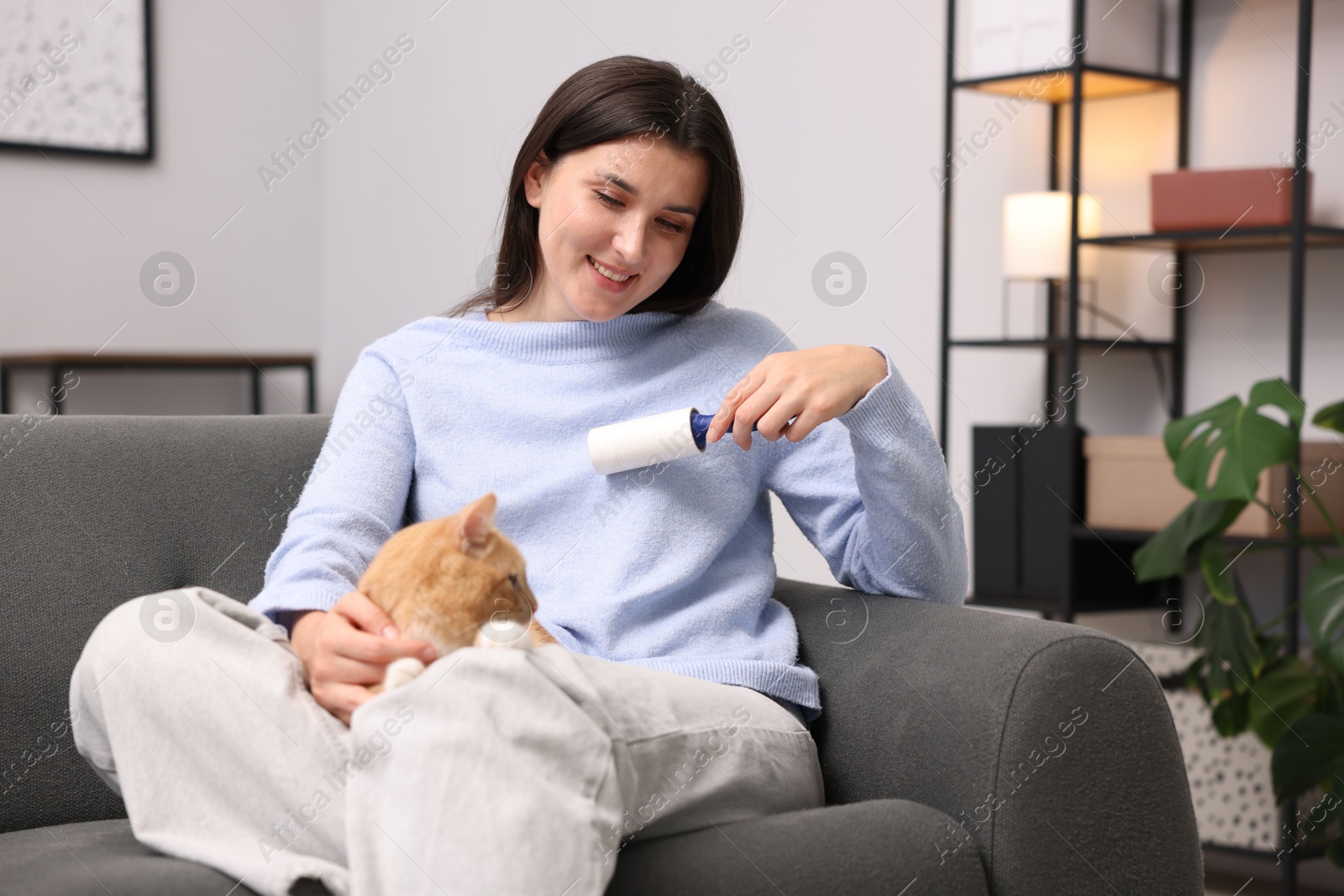 Photo of Smiling woman with lint roller removing pet's hair from clothes and her cat on sofa at home