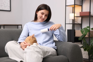 Photo of Smiling woman with lint roller removing pet's hair from clothes and her cat on sofa at home