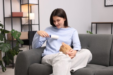 Photo of Smiling woman with lint roller removing pet's hair from clothes and her cat on sofa at home