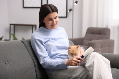 Photo of Smiling woman with lint roller removing pet's hair from clothes and her cat on sofa at home