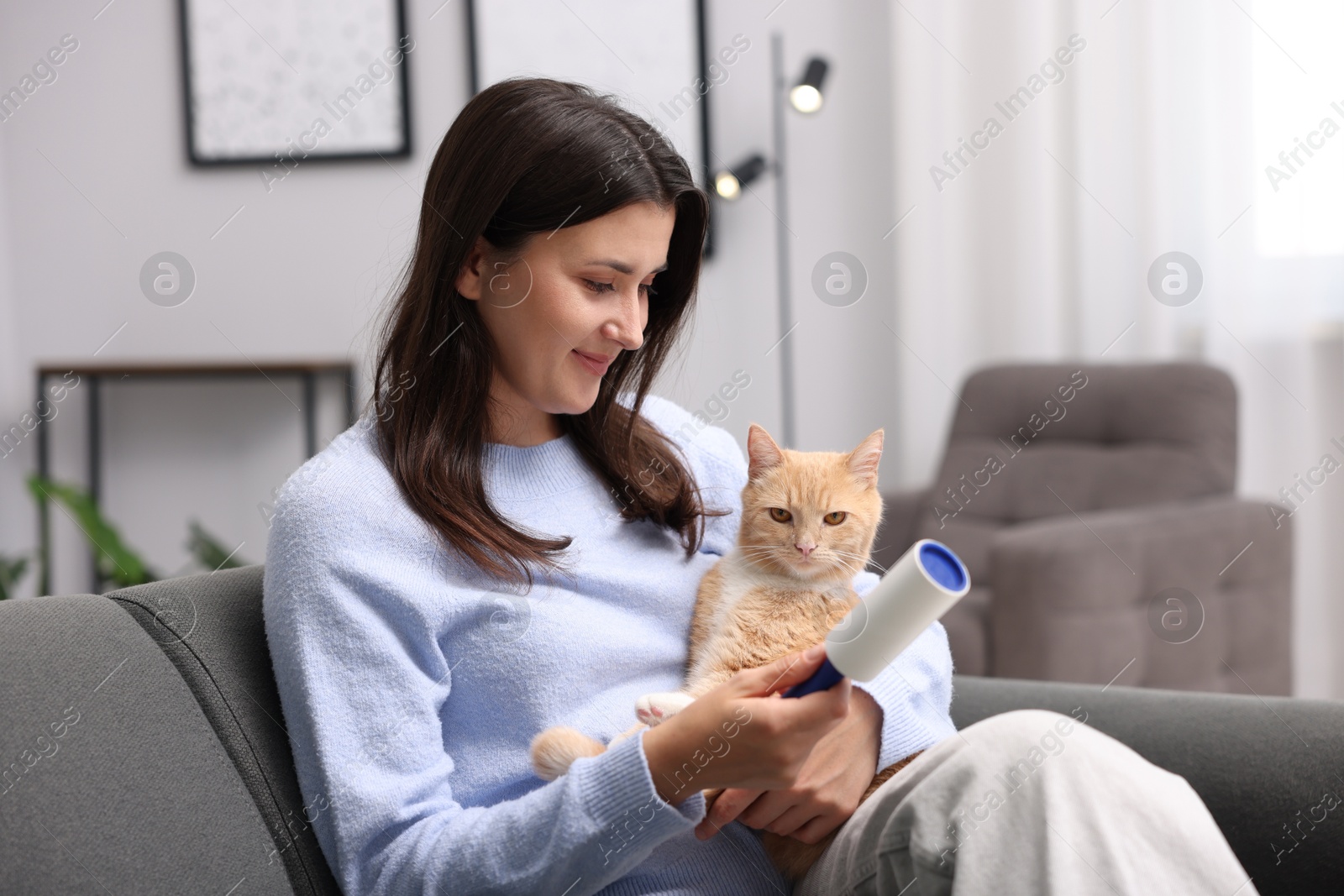 Photo of Smiling woman with lint roller and her cat on sofa at home