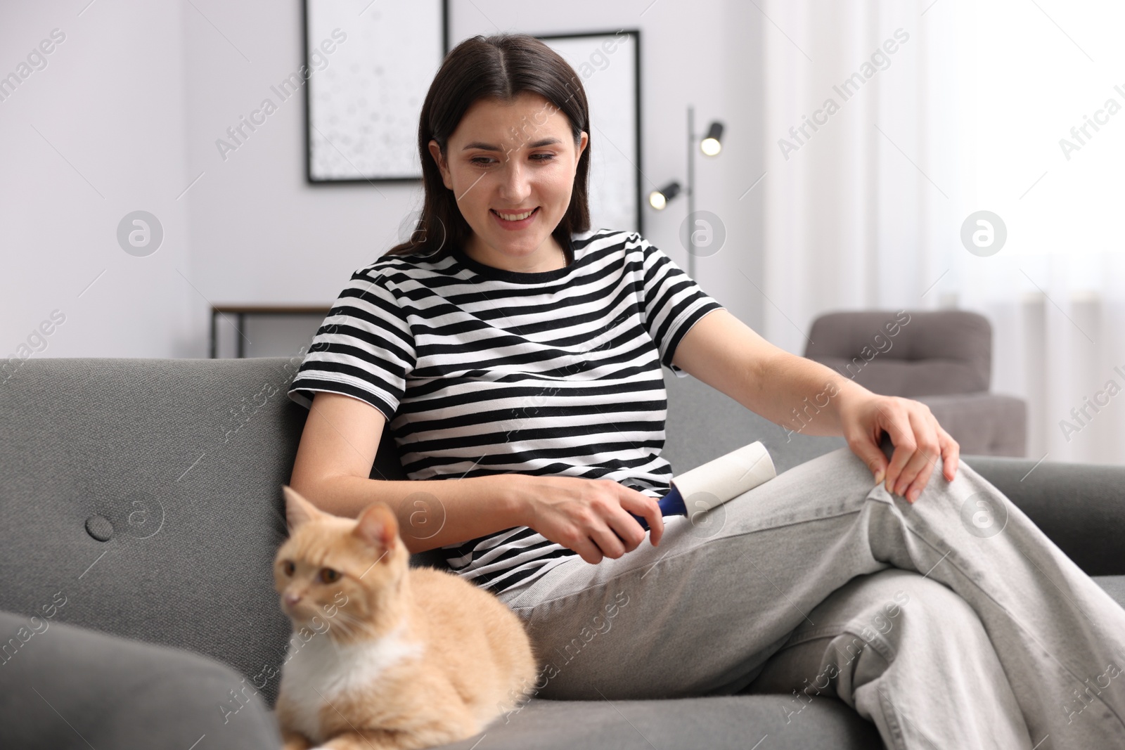 Photo of Smiling woman with lint roller removing pet's hair from clothes and her cat on sofa at home