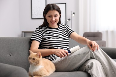 Photo of Smiling woman with lint roller removing pet's hair from clothes and her cat on sofa at home